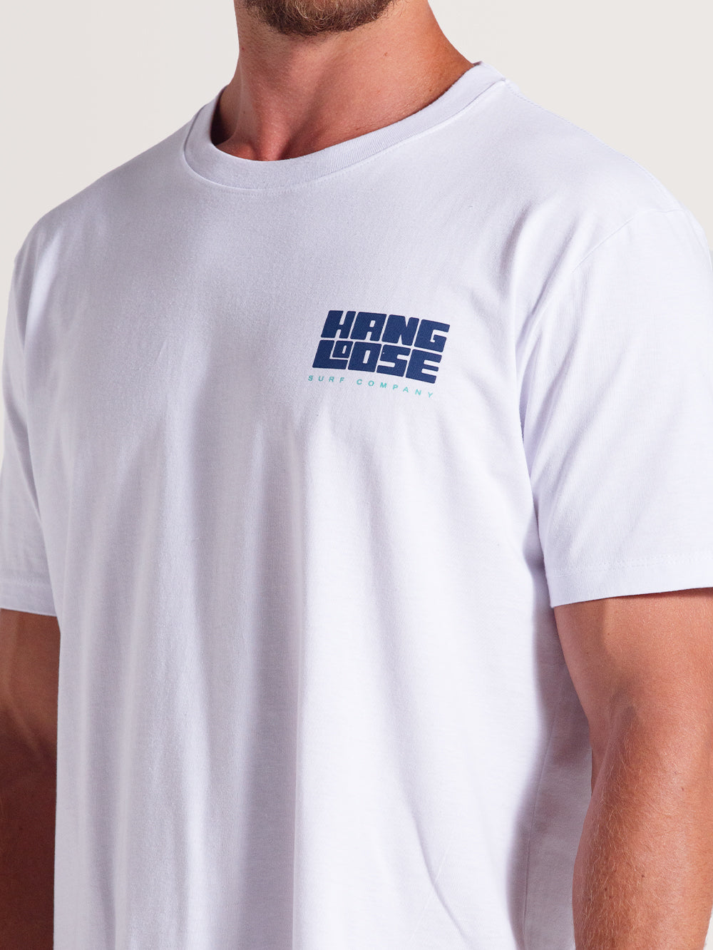Homem vestindo camiseta branca manga curta com logo azul "Hang Loose Surf Company" no lado esquerdo do peito. Estilo casual.
