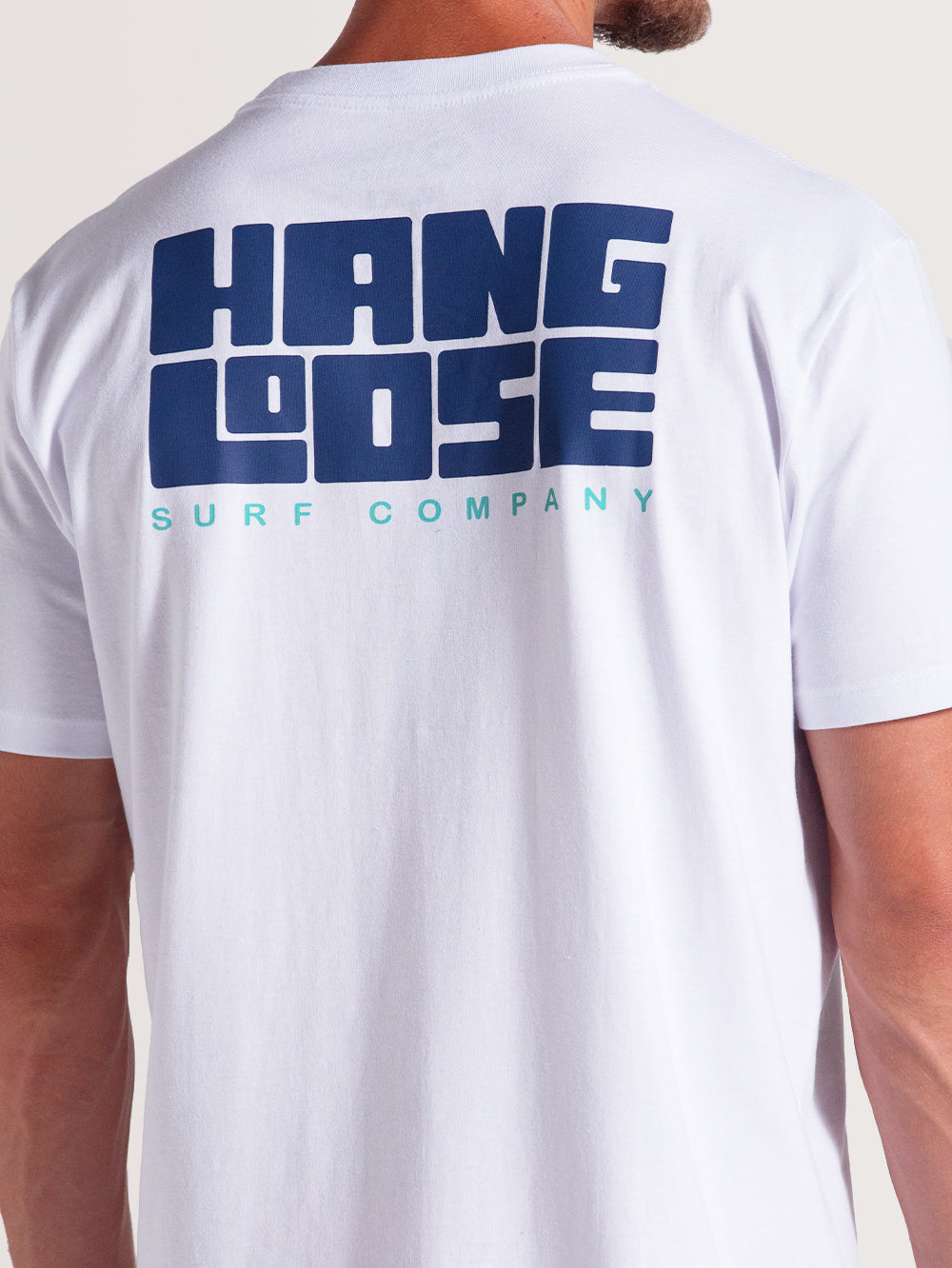 Homem vestindo camiseta branca manga curta com estampa traseira grande em azul escuro "Hang Loose" e texto menor "Surf Company" em azul claro.