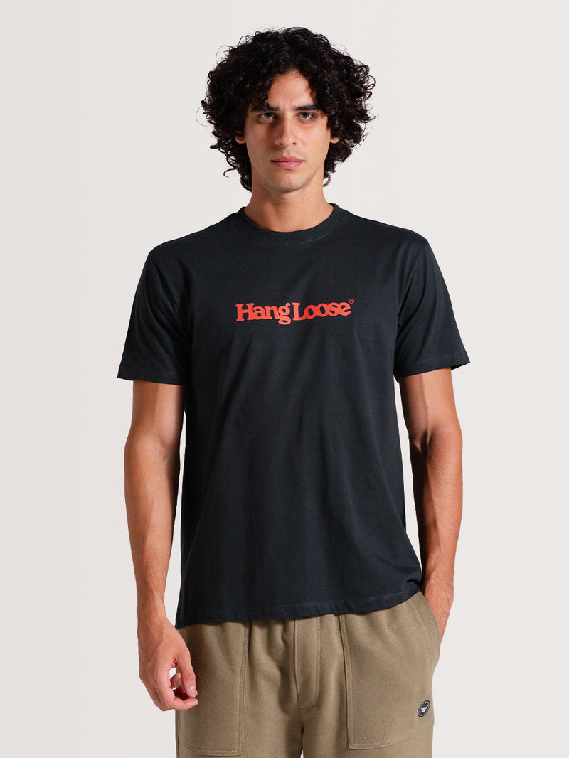 Homem vestindo camiseta cinza algodão manga curta com gola redonda e logo "Hang Loose" vermelho no peito, combinada com calça caqui, sobre fundo claro.