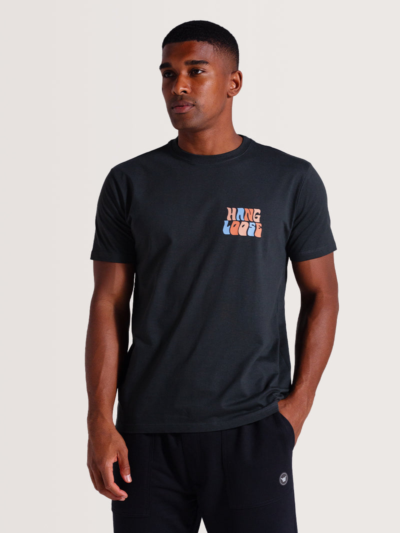 Homem vestindo camiseta cinza básica manga curta com estampa "Hang Loose" estilizada em azul e laranja no lado esquerdo do peito, combinada com calça preta casual.