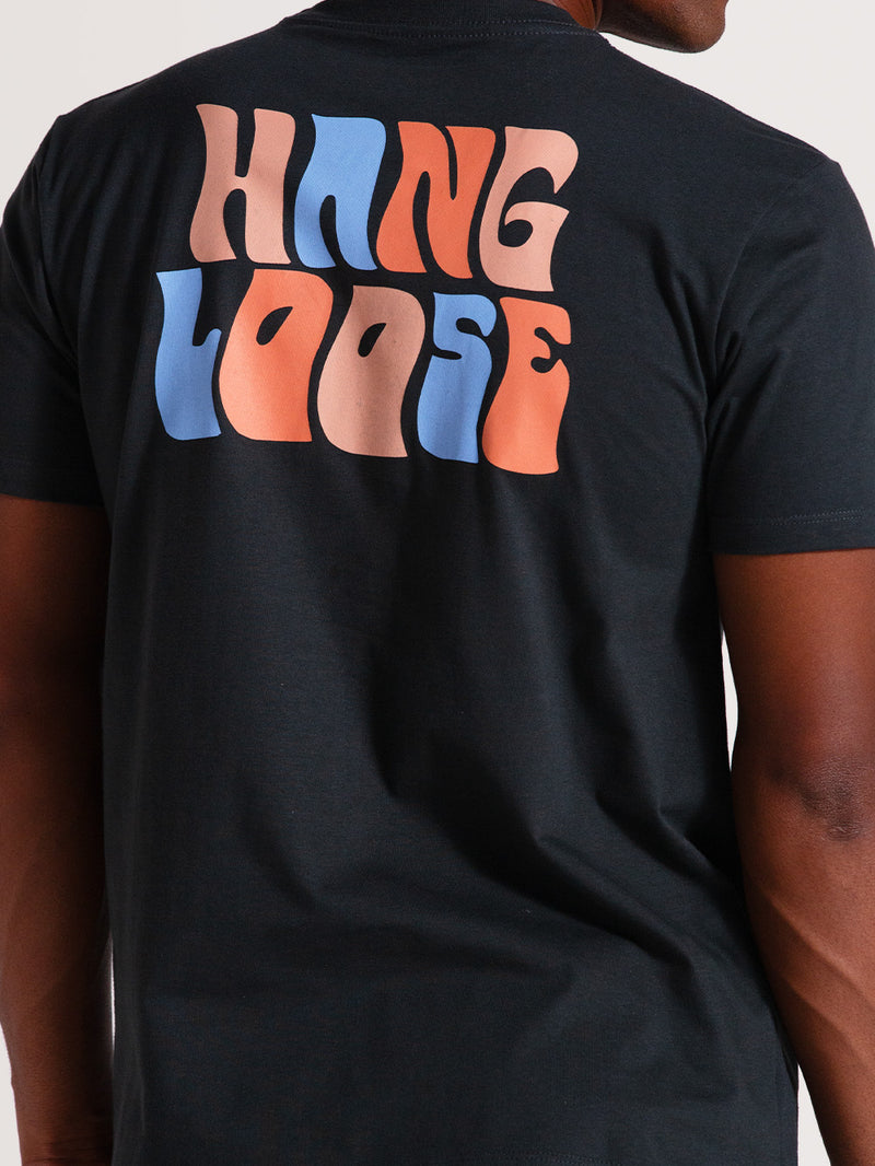 Homem vestindo uma camiseta cinza básica manga curta com estampa traseira "Hang Loose" estilizada em laranja, azul e bege em um design vibrante.