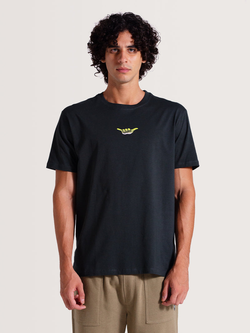 Homem vestindo camiseta cinza chumbo manga curta com pequeno símbolo "Hang Loose" verde-limão no centro do peito, combinada com calça bege.