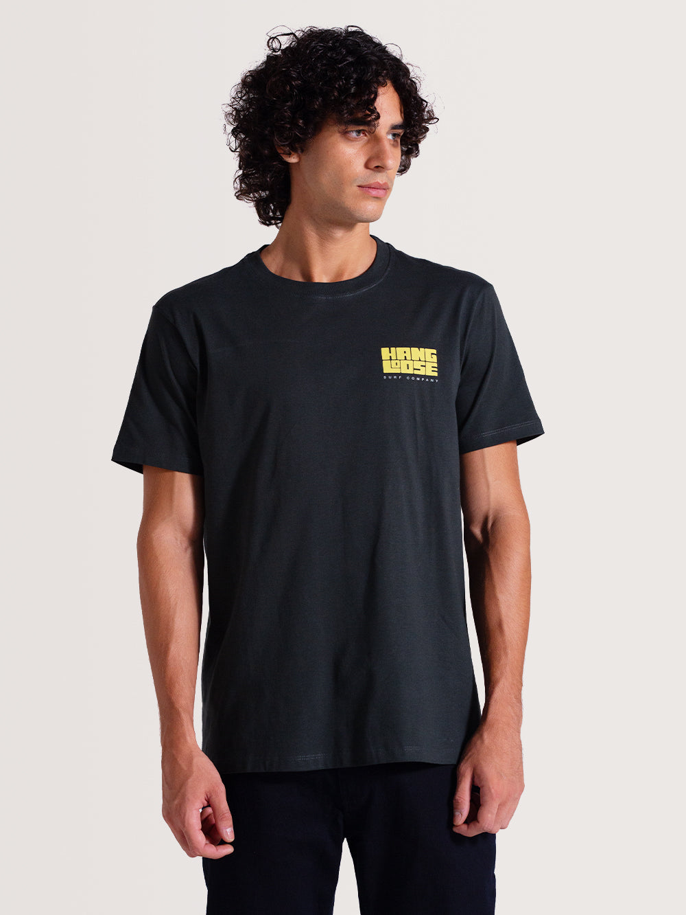 Homem vestindo camiseta cinza manga curta com pequeno logotipo amarelo "Hang Loose Surf Company" no lado esquerdo do peito. Estilo casual.