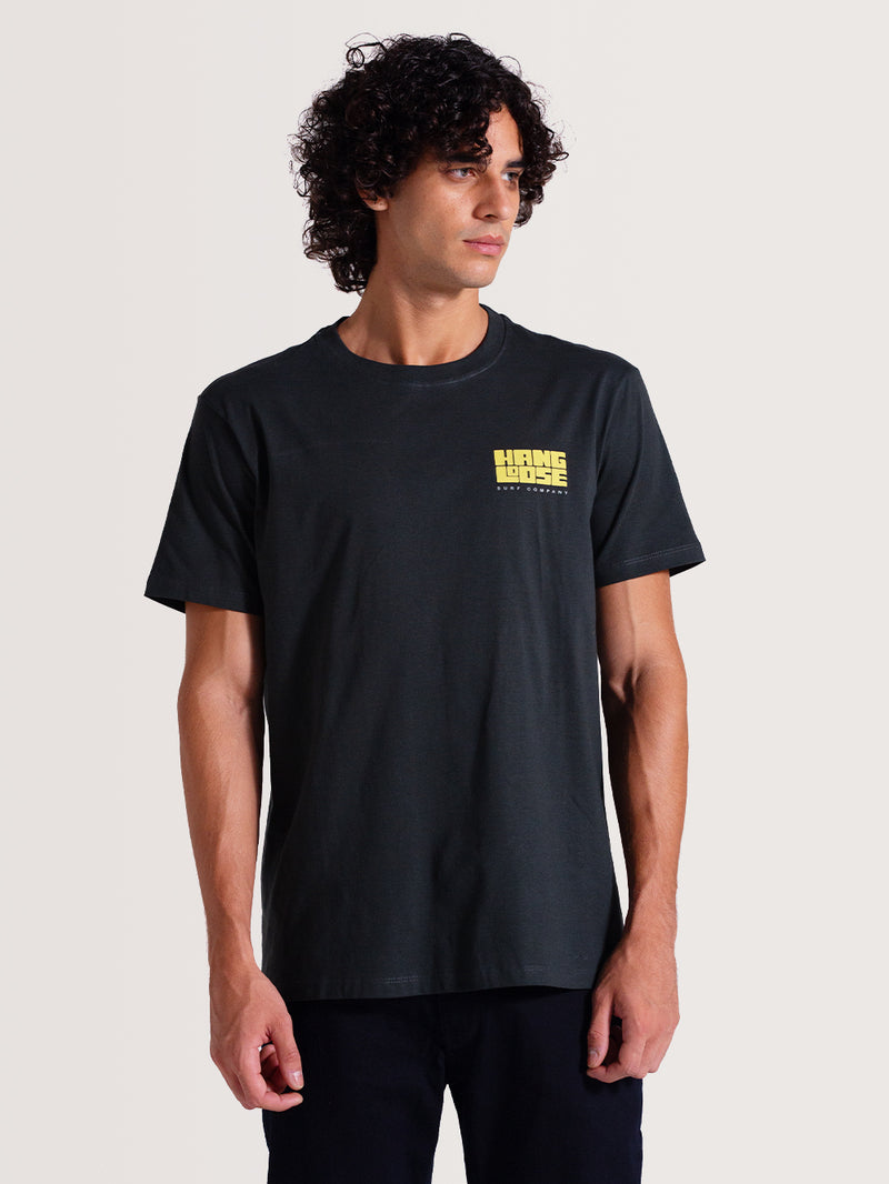 Homem vestindo camiseta cinza manga curta com pequeno logotipo amarelo "Hang Loose Surf Company" no lado esquerdo do peito. Estilo casual.
