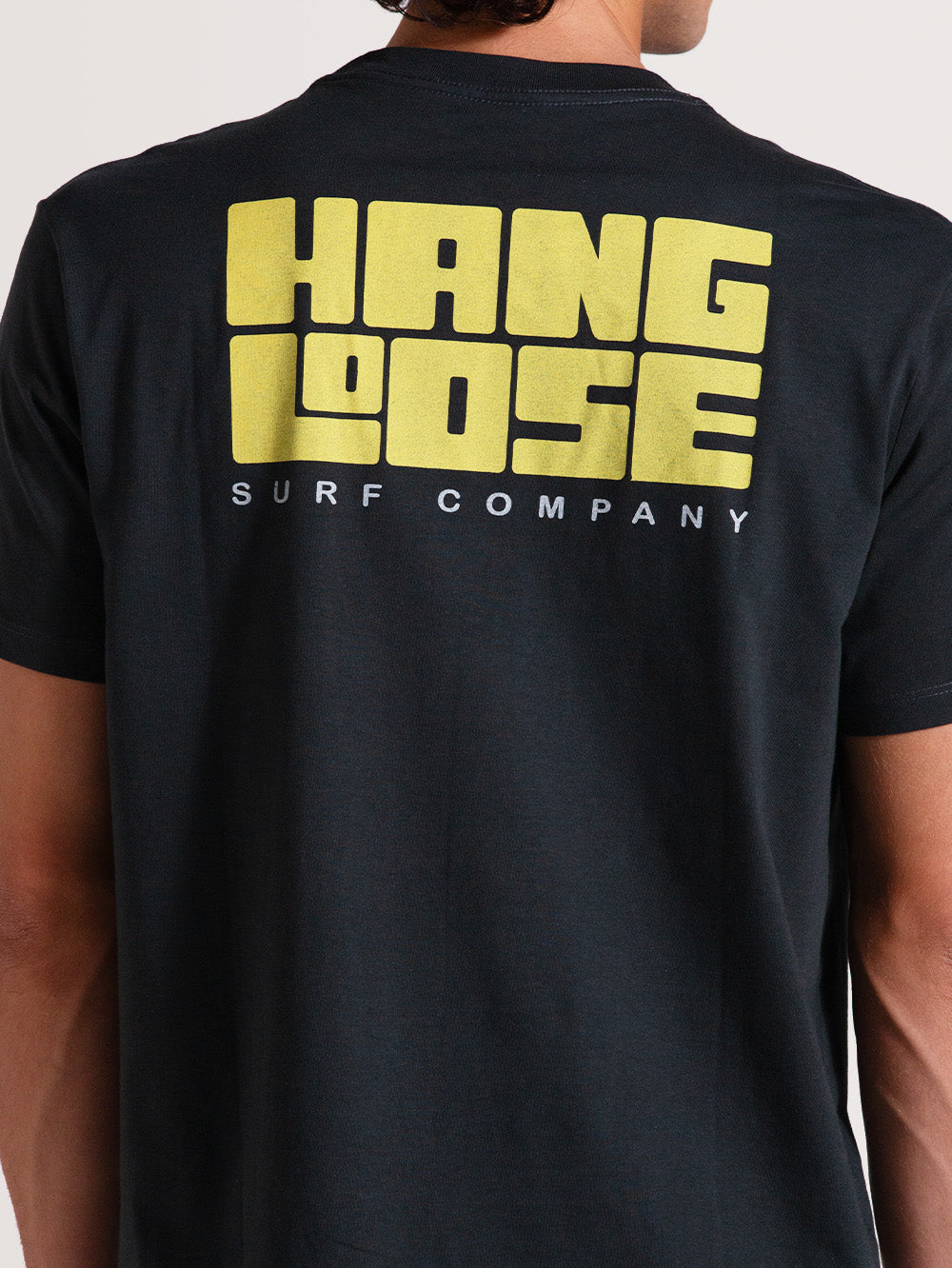 Homem vestindo camiseta cinza manga curta com estampa traseira grande em amarelo "Hang Loose Surf Company". Estilo casual e moderno.