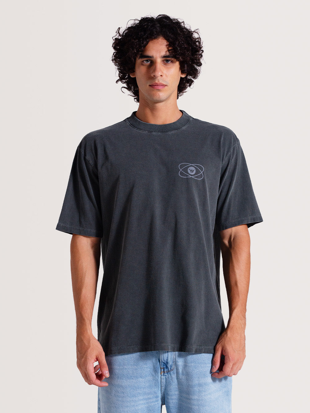 Homem usando camiseta cinza manga longa com design minimalista no peito e tecido em tom desbotado, combinada com jeans claro, em fundo neutro.