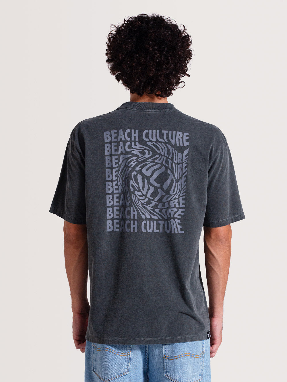 Homem de costas vestindo camiseta cinza manga longa com estampa nas costas escrita "BEACH CULTURE" repetida em azul, formando um efeito ondulado, em fundo claro.