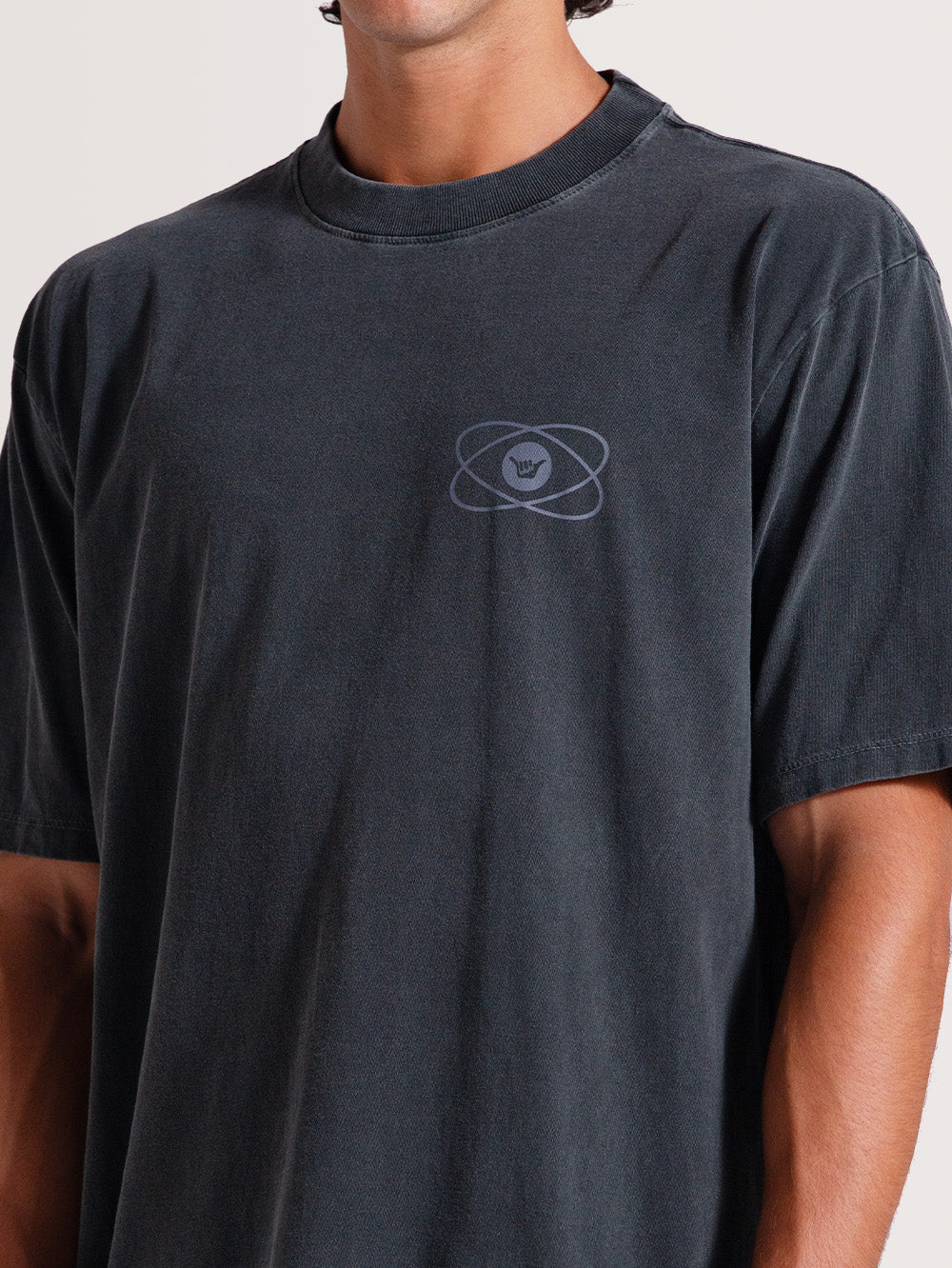 Homem vestindo camiseta cinza manga longa com símbolo minimalista no peito de círculos entrelaçados e o gesto "shaka" ao centro, sobre fundo claro.