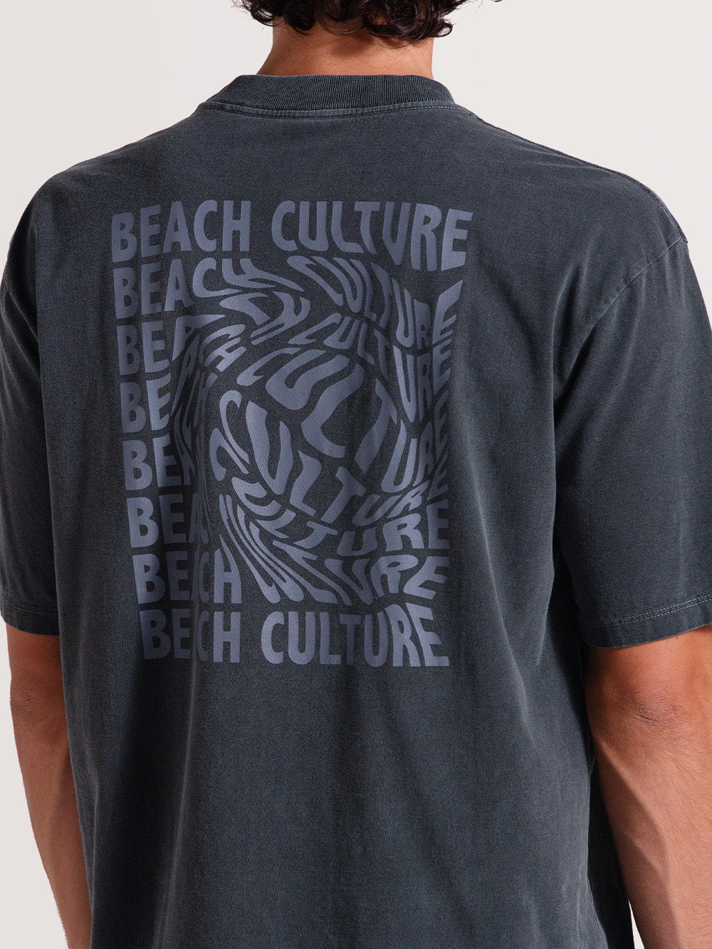 Homem de costas vestindo camiseta cinza manga longa com estampa "BEACH CULTURE" repetida em tom mais claro, criando efeito ondulado, sobre tecido desbotado.