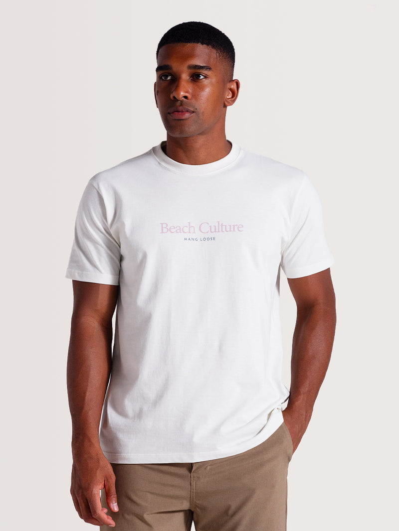 Homem vestindo camiseta cor off white manga curta com estampa minimalista rosa claro escrita "Beach Culture Hang Loose" no centro do peito.