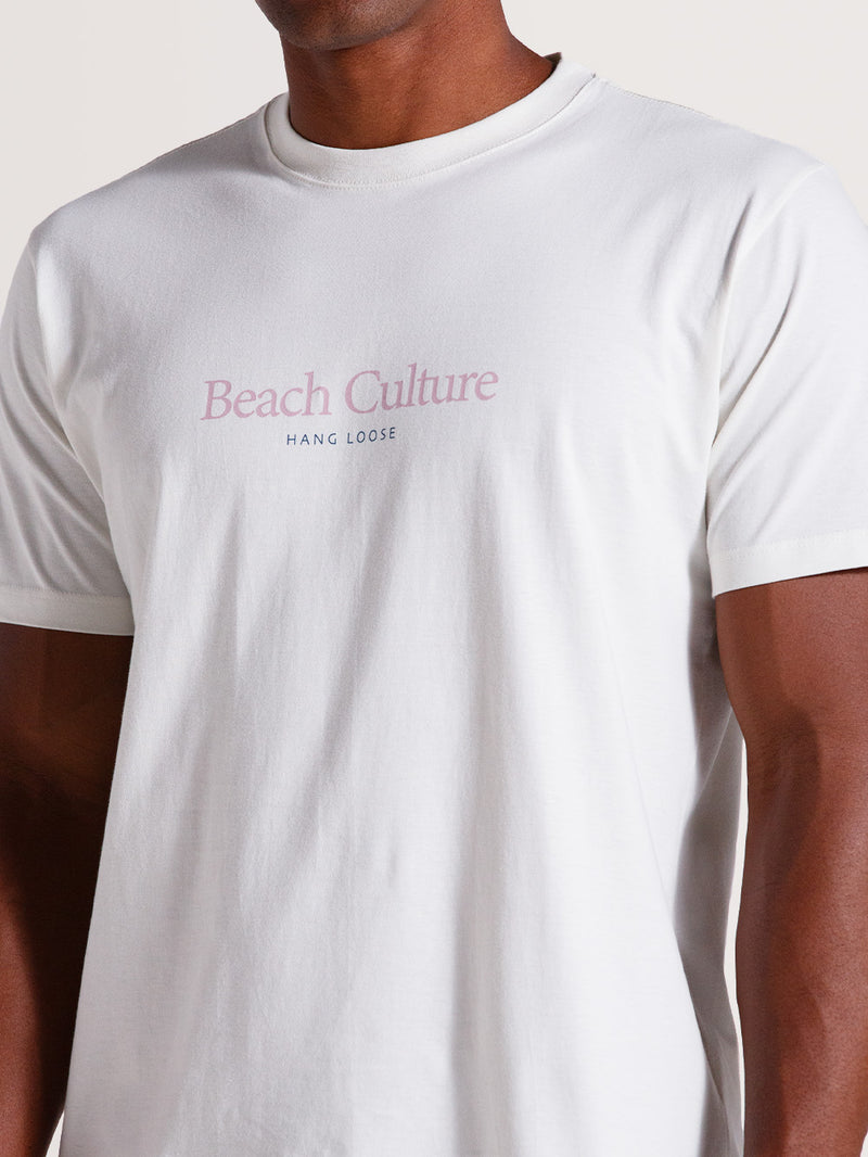 Homem vestindo camiseta cor off white manga curta com a frase "Beach Culture Hang Loose" estampada no centro do peito em tons rosa e azul.