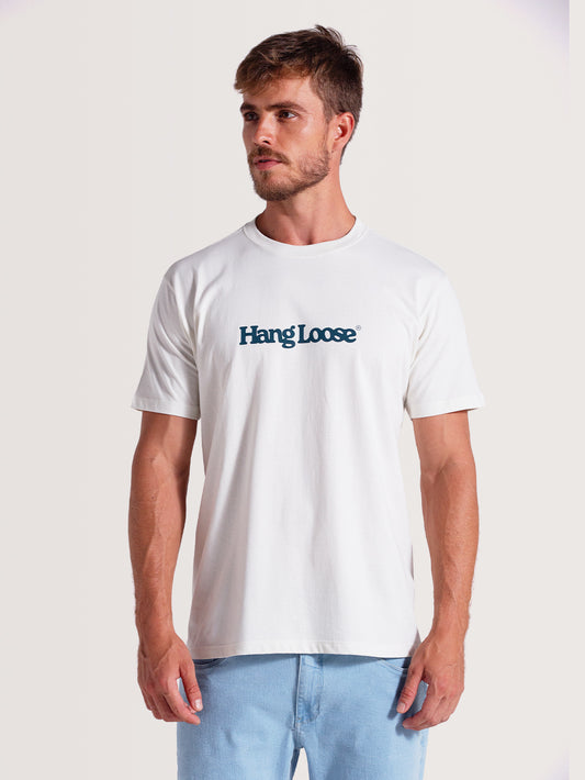 Homem vestindo camiseta off white algodão manga curta com gola redonda e logo "Hang Loose" azul no peito, combinada com calça jeans azul claro, sobre fundo neutro.