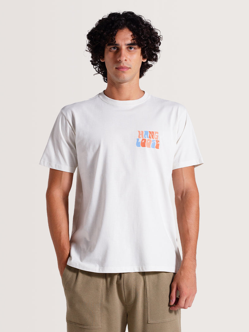 Homem vestindo camiseta off white básica manga curta com estampa "Hang Loose" em azul e laranja no lado esquerdo do peito, combinada com calça bege.