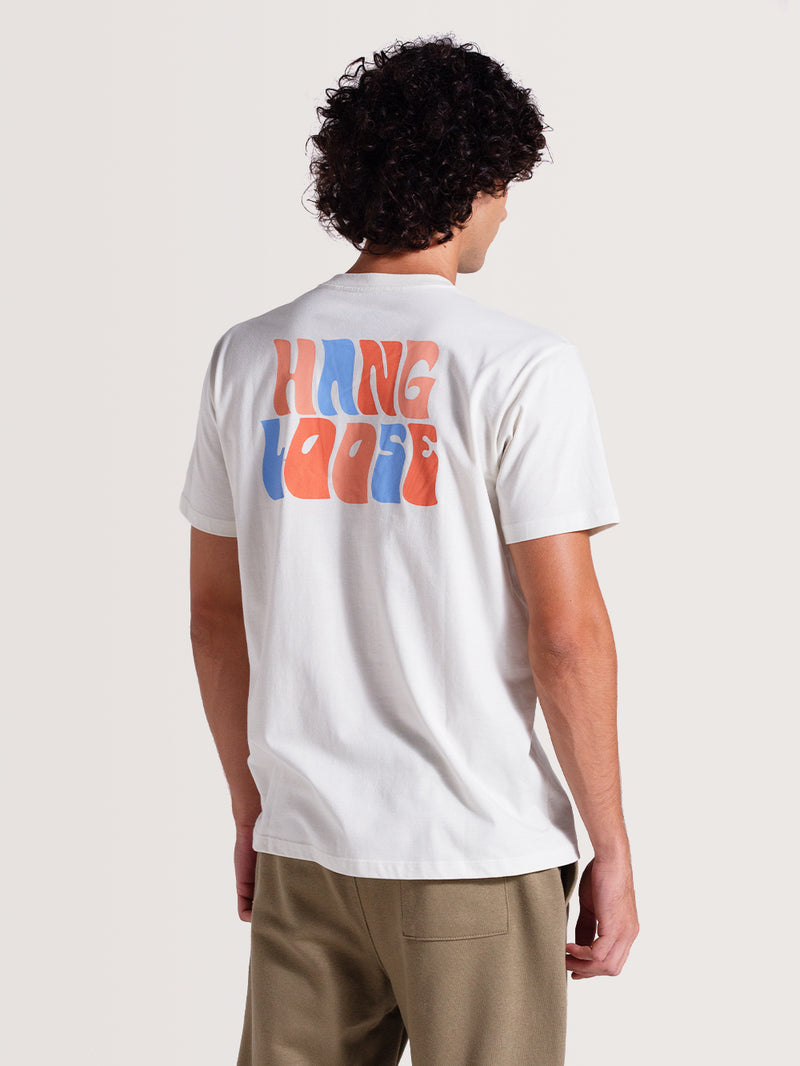 Homem vestindo uma camiseta off white básica manga curta com estampa traseira "Hang Loose" em letras grandes nas cores azul e laranja vibrantes.