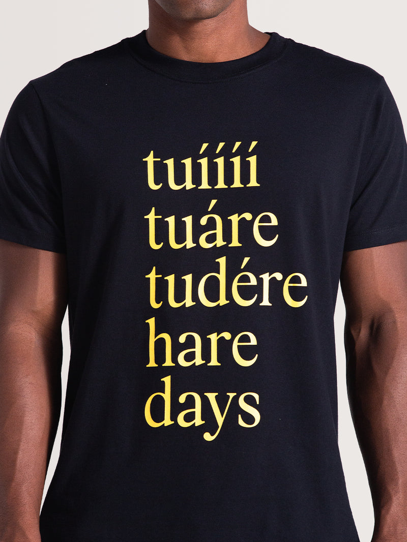 Homem vestindo uma camiseta preta de manga curta com estampa criativa em amarelo. A frase “tuíííí, tuáre, tudére, hare days” está centralizada e em destaque.