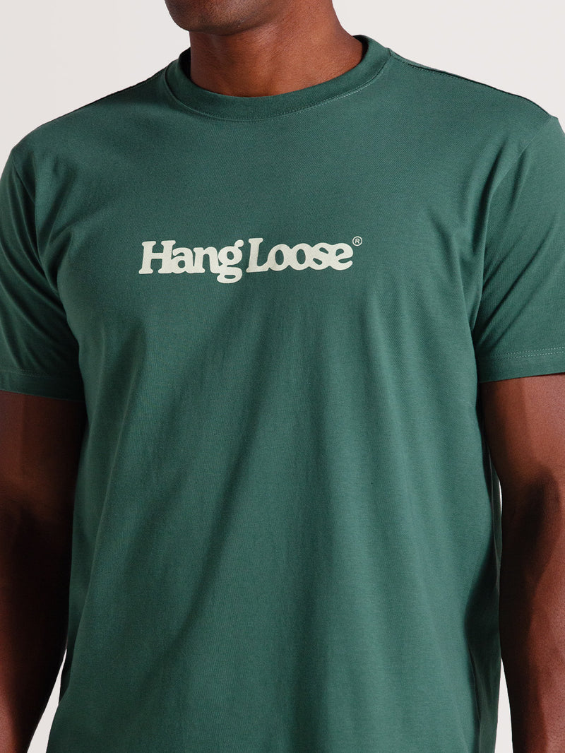Homem vestindo camiseta verde algodão manga curta com gola redonda e logo "Hang Loose" branco no peito, em fundo claro.