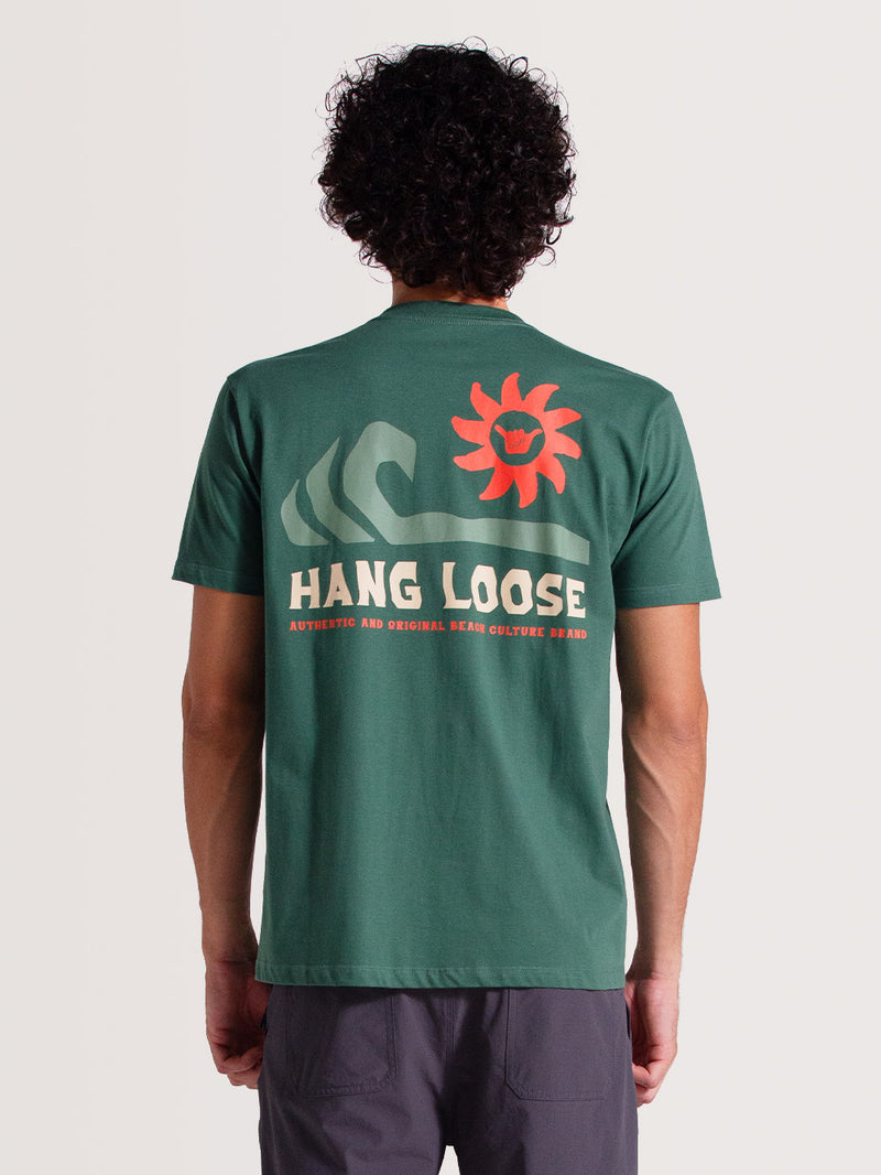 Homem de costas vestindo camiseta verde musgo manga curta com estampa grande de ondas, sol e texto "Hang Loose" em laranja e branco, em fundo claro.