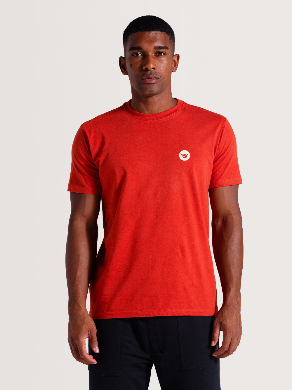 Homem vestindo camiseta vermelha manga curta lisa, com pequeno logo branco no lado esquerdo do peito, combinada com calça preta. Estilo minimalista.