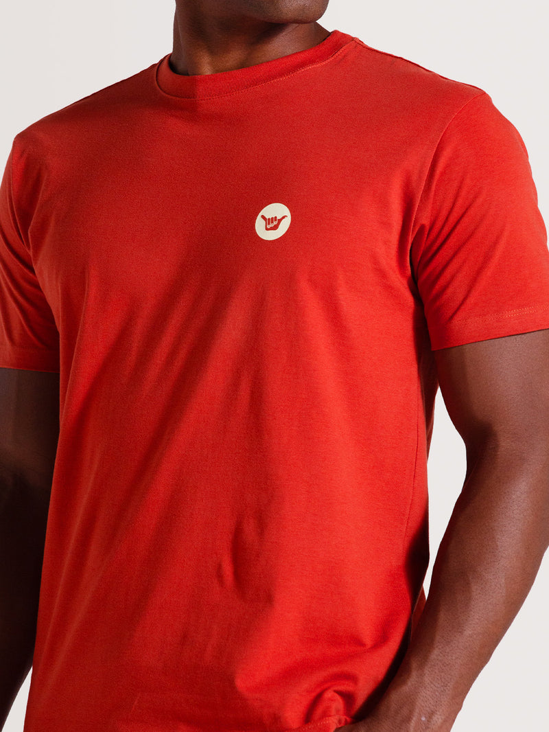 Camiseta vermelha manga curta lisa com um pequeno logo branco no peito, representando o gesto "hang loose". Estilo casual e clean.
