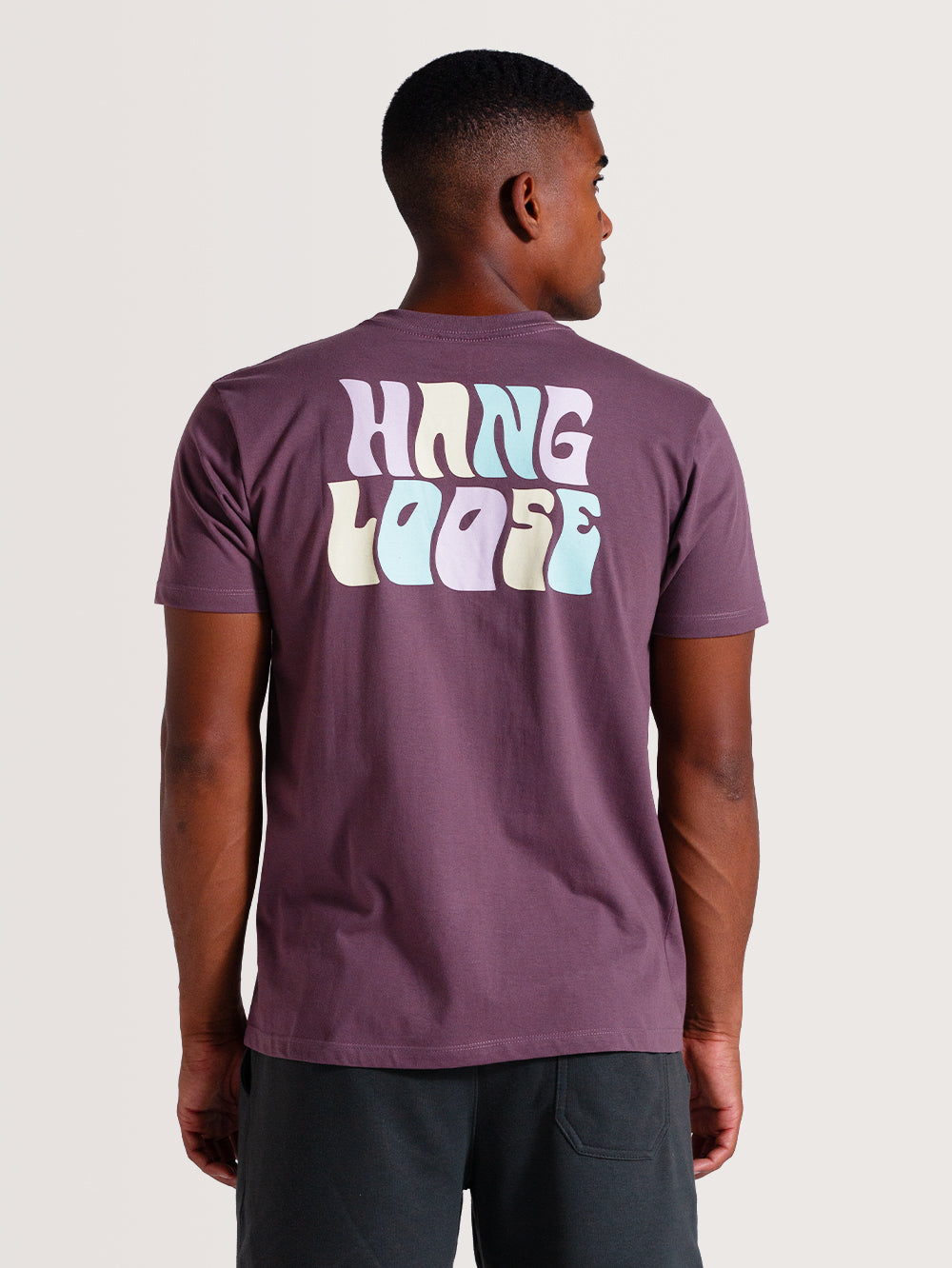 Homem vestindo camiseta violeta manga curta com estampa "Hang Loose" colorida nas costas, combinada com shorts escuros.