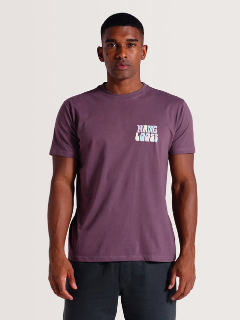 Homem vestindo camiseta violeta manga curta com estampa frontal "Hang Loose" branca estilizada, combinada com shorts preto básico.