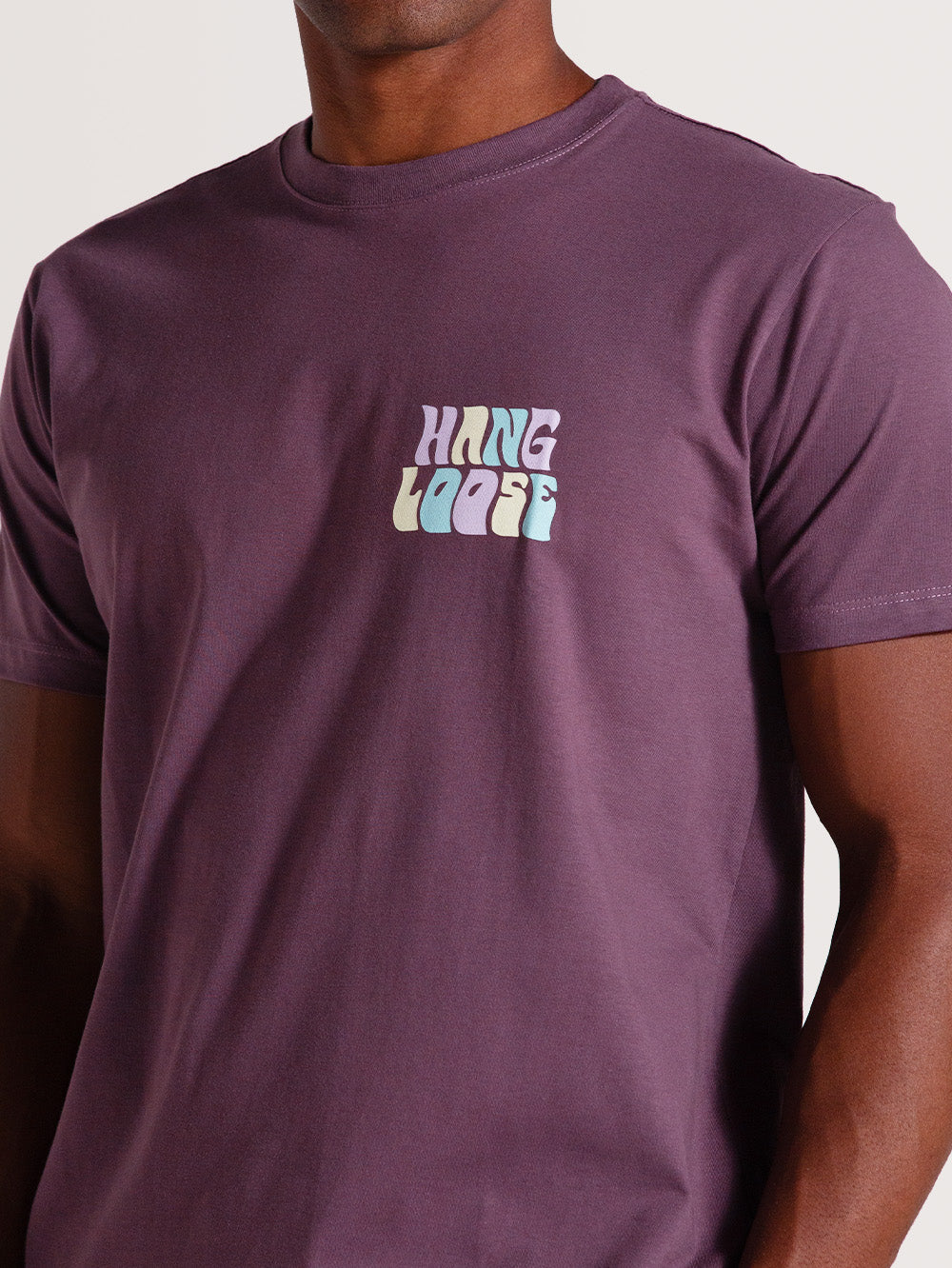 Homem usando camiseta violeta manga curta com estampa frontal "Hang Loose" em tons pastel, sobre fundo neutro.