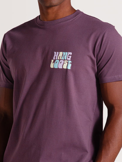 Homem usando camiseta violeta manga curta com estampa frontal "Hang Loose" em tons pastel, sobre fundo neutro.