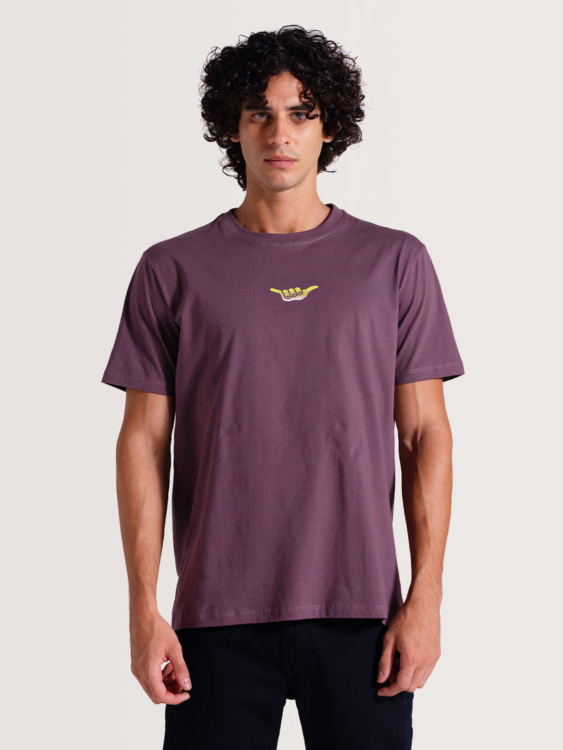 Homem vestindo uma camiseta violeta manga curta com pequeno símbolo estilizado "Hang Loose" em amarelo no centro do peito, combinada com calça escura.