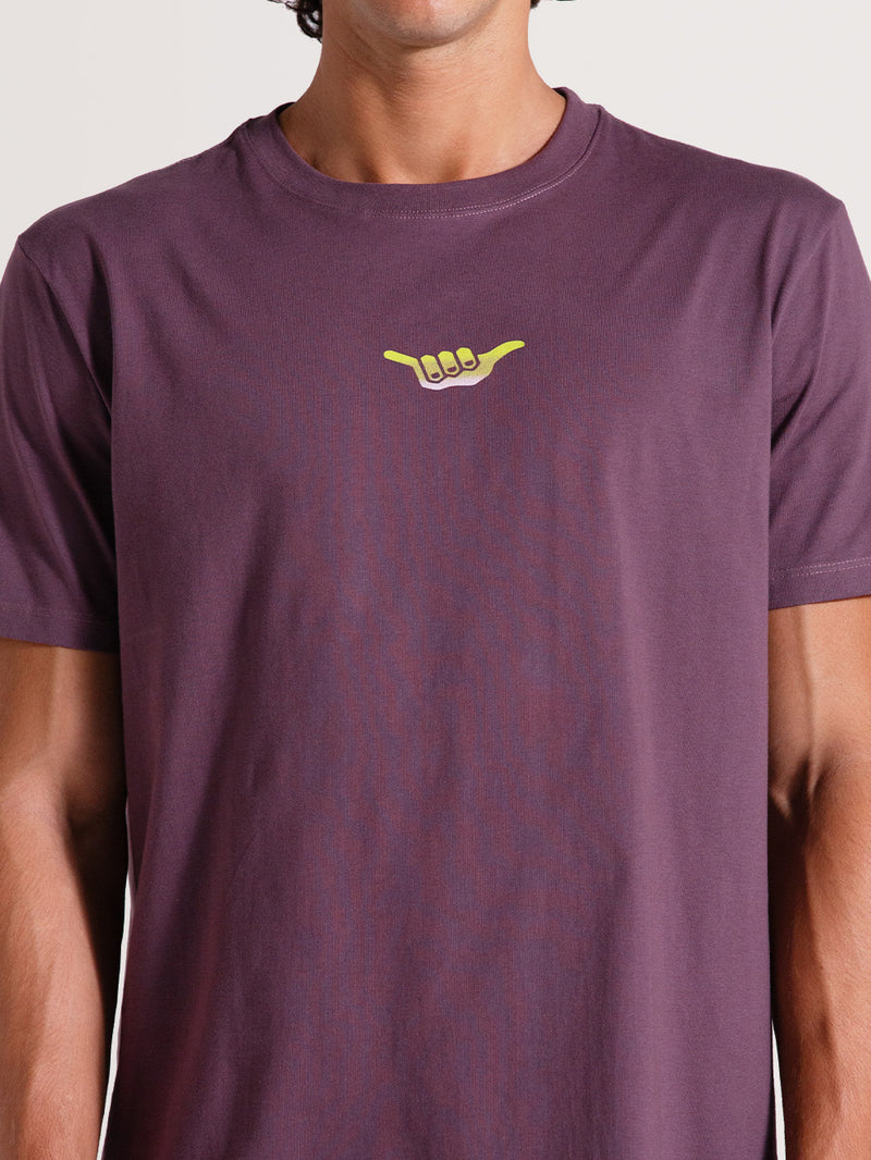 Homem vestindo camiseta violeta manga curta com estampa frontal de uma mão fazendo o gesto "Hang Loose" em amarelo vibrante no centro do peito.