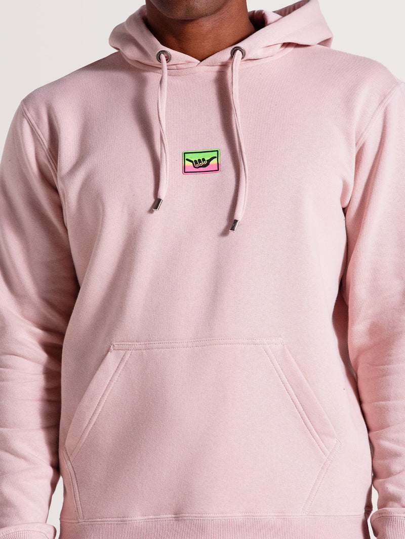 Close do moletom canguru rosa com capuz, bolso frontal e cordões ajustáveis. Destaque para patch colorido com símbolo de mão no peito.