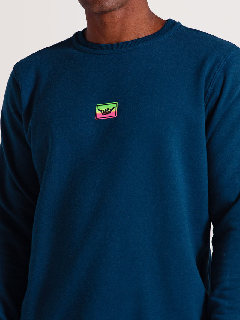 Moletom careca azul em tom escuro, com logo colorido em verde, rosa e amarelo no peito. Corte reto, acabamento simples nos punhos e gola para estilo casual.