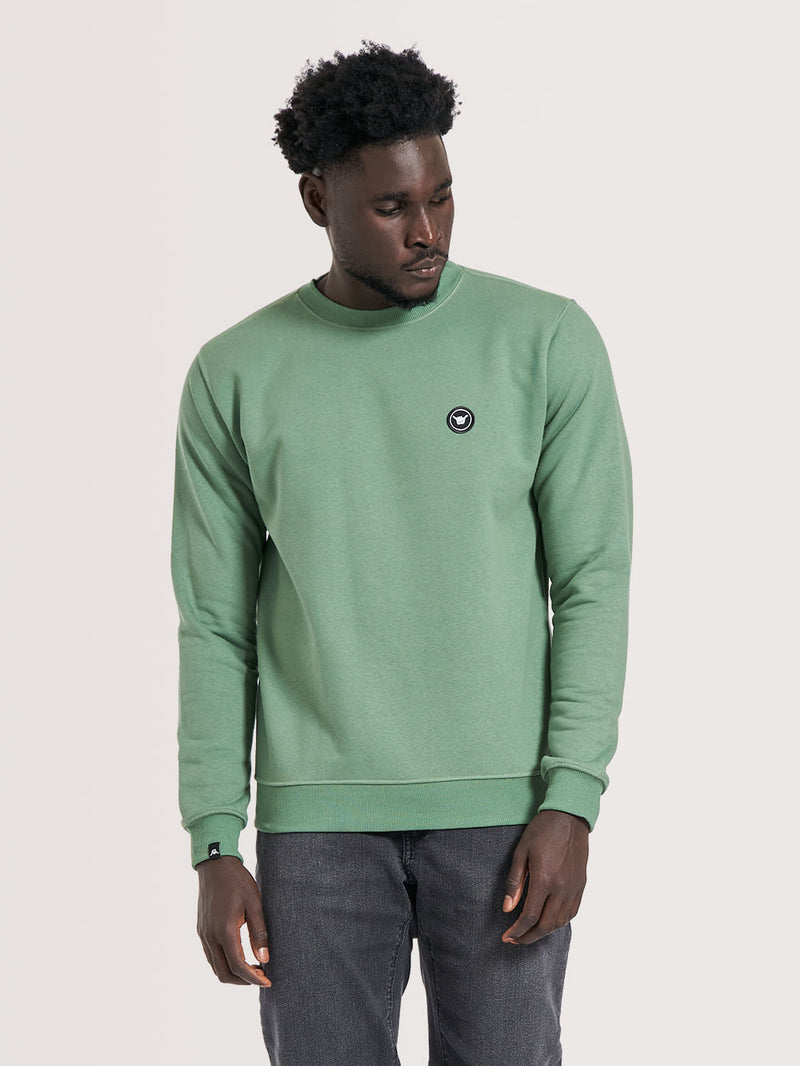 Pessoa vestindo moletom careca verde com gola redonda, detalhe de emblema preto no peito e modelagem simples. Combina com calça jeans escura.