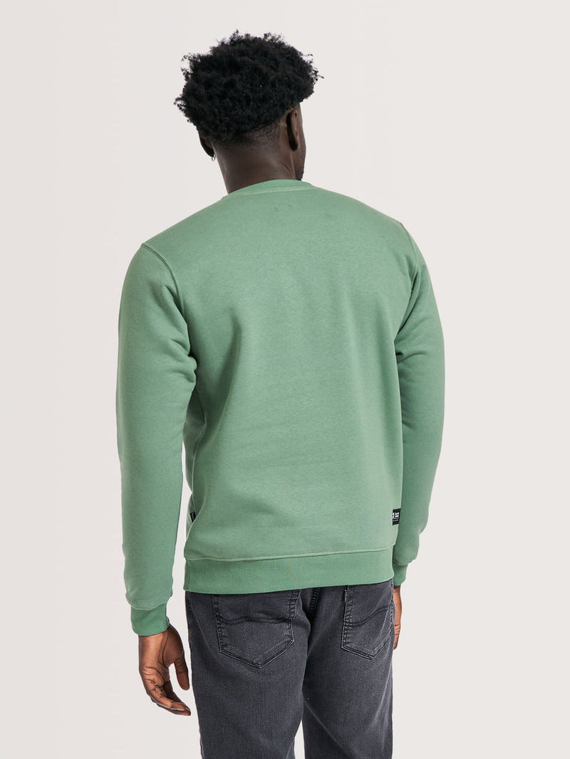 Homem de costas vestindo moletom careca verde, de modelagem simples, gola redonda e tecido liso. Combina com jeans escuro casual.
