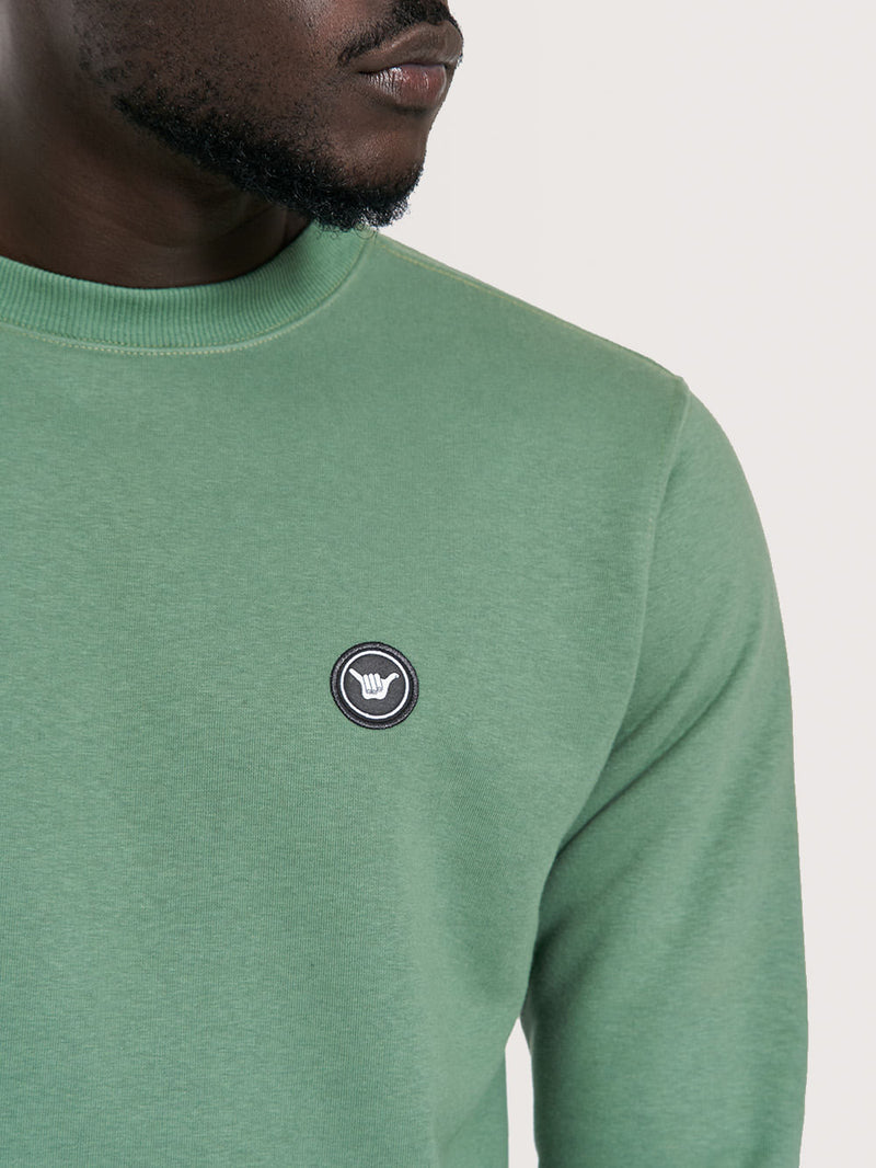 Homem usando moletom careca verde com gola redonda e emblema circular bordado no peito esquerdo.