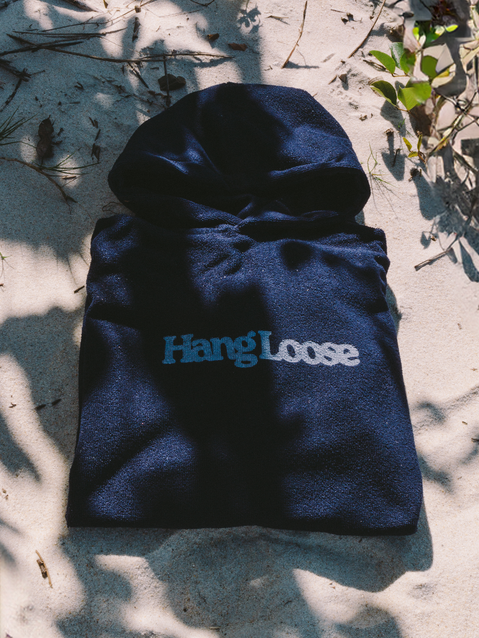 Poncho surf azul marinho com capuz e logo da Hang Loose dobrado sobre a areia, destacando estilo e praticidade para dias de praia.