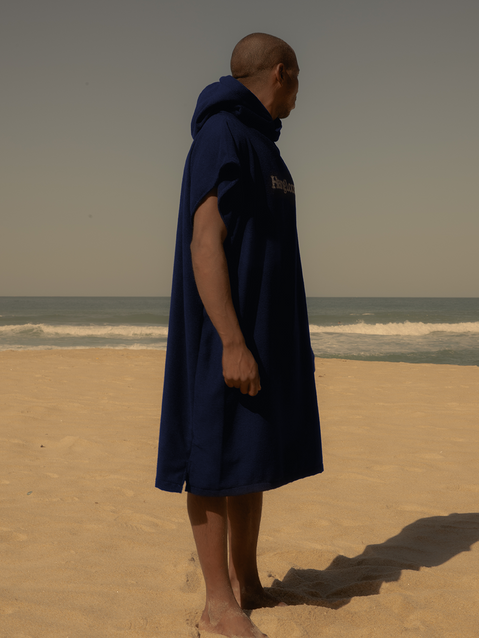 Homem de costas na praia vestindo poncho surf azul marinho com capuz da Hang Loose, perfeito para praticantes de surfe e atividades à beira-mar.