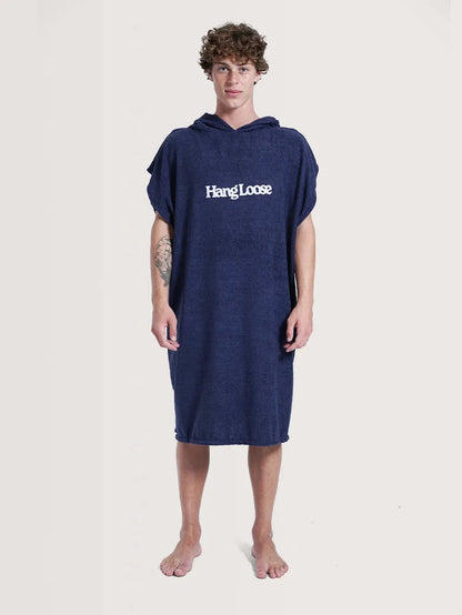 Modelo posando em fundo neutro vestindo o poncho surf da Hang Loose. A peça possui cor azul e nome da marca em branco na parte frontal.