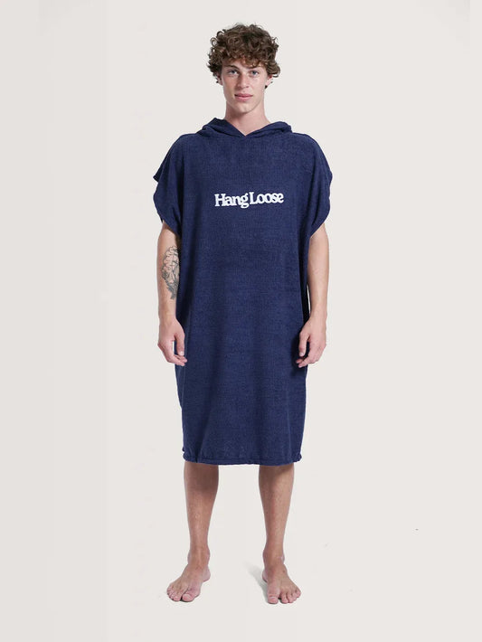 Modelo posando em fundo neutro vestindo o poncho surf da Hang Loose. A peça possui cor azul e nome da marca em branco na parte frontal.