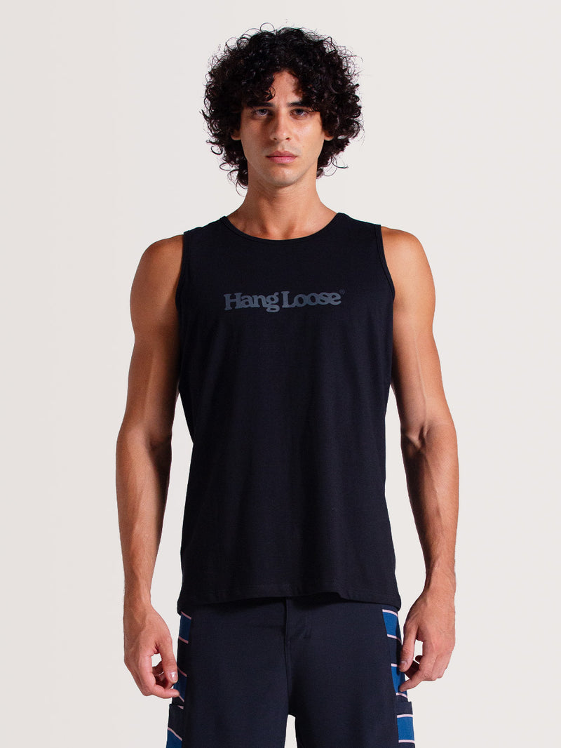 Homem vestindo regata preta básica com logo "Hang Loose" estampado em cinza no peito, combinada com bermuda azul com detalhes coloridos, em fundo claro.
